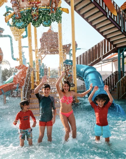 Four young children playing in shallow water at a splash pad with spraying water features and blue tube slides
