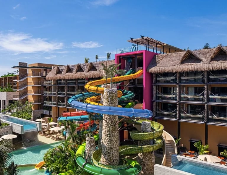 Parque acuático tropical con coloridos toboganes en espiral, palmeras, albercas y edificios con techo de paja bajo el cielo azul