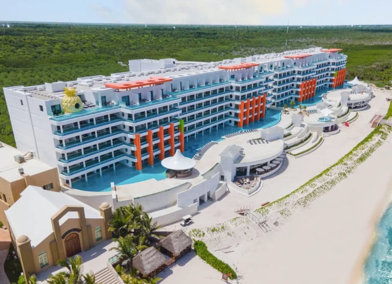 Vista aérea de un resort moderno frente al mar con arquitectura azul y naranja, albercas y vegetación tropical