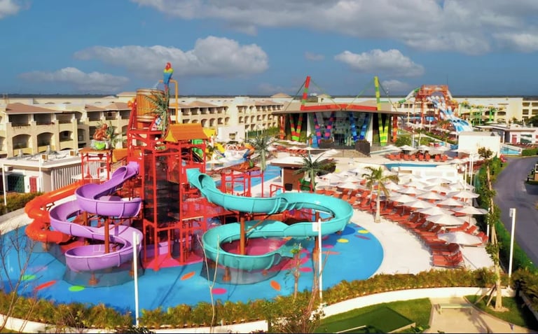 Colorful water park with purple and blue slide towers, wave pools, and attractions surrounded by blue flooring on a sunny day