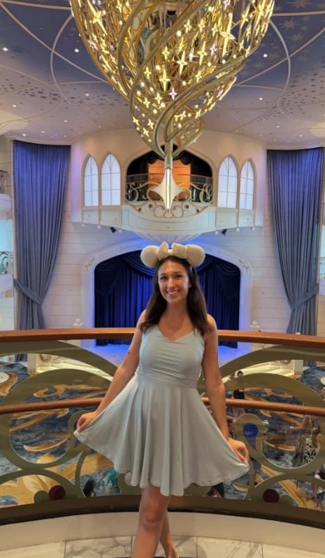 Woman in light blue dress and mouse ears posing in luxurious hallway with ornate chandelier and blue curtains